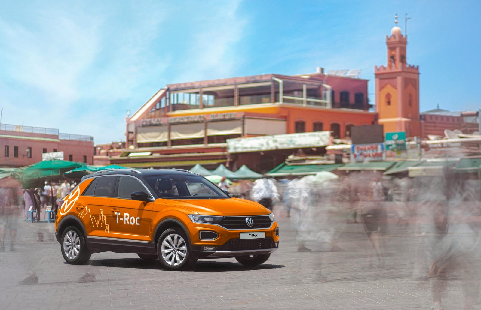 Orange SUV in motion at a busy Marrakech market. Vibrant urban scene with traditional architecture.