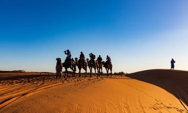 A group of travelers on camels crossing the sandy dunes of the Sahara Desert under a clear blue sky.